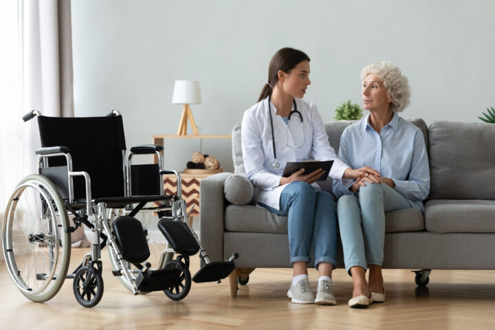 A doctor sits next to an elderly woman on a sofa and holds her hand. There is a wheelchair to the right of the sofa.