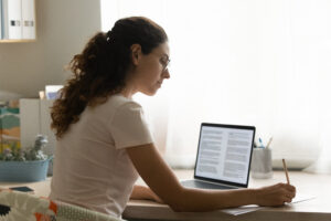 A woman sitting at a desk with an open laptop and notebook in front of her, she is looking down and writing notes.