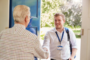 An elderly man with his back to the camera opens his front door to another man, who is wearing a lanyard.